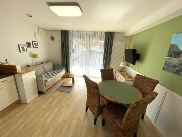 Une pièce moderne et bien éclairée, avec un canapé, une table à manger, des chaises en osier, un téléviseur et un tableau accroché au mur.