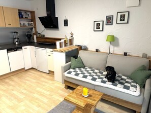 Un espace de vie compact comprenant un coin cuisine, un canapé avec un chien, une petite table avec une bougie et des tableaux encadrés accrochés au mur.