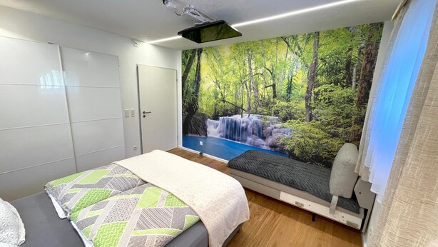 Une chambre à coucher comprenant un lit, un projecteur fixé au mur, une armoire et une fresque murale représentant une cascade dans une forêt.