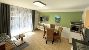Un salon comprenant un canapé, une table à manger avec des chaises, un téléviseur fixé au mur et un coin cuisine équipé d’un four à micro-ondes et d’un four traditionnel.