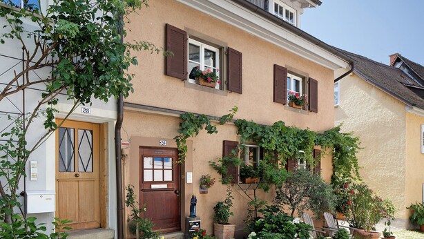 A two-story building with a beige facade, wooden shutters, and a small garden with potted plants and flowers. - Apartment "Gill" with Wi-Fi (Staufen)