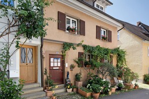 Een gebouw met twee verdiepingen, een beige gevel, houten luiken en een kleine tuin met potplanten en bloemen.
