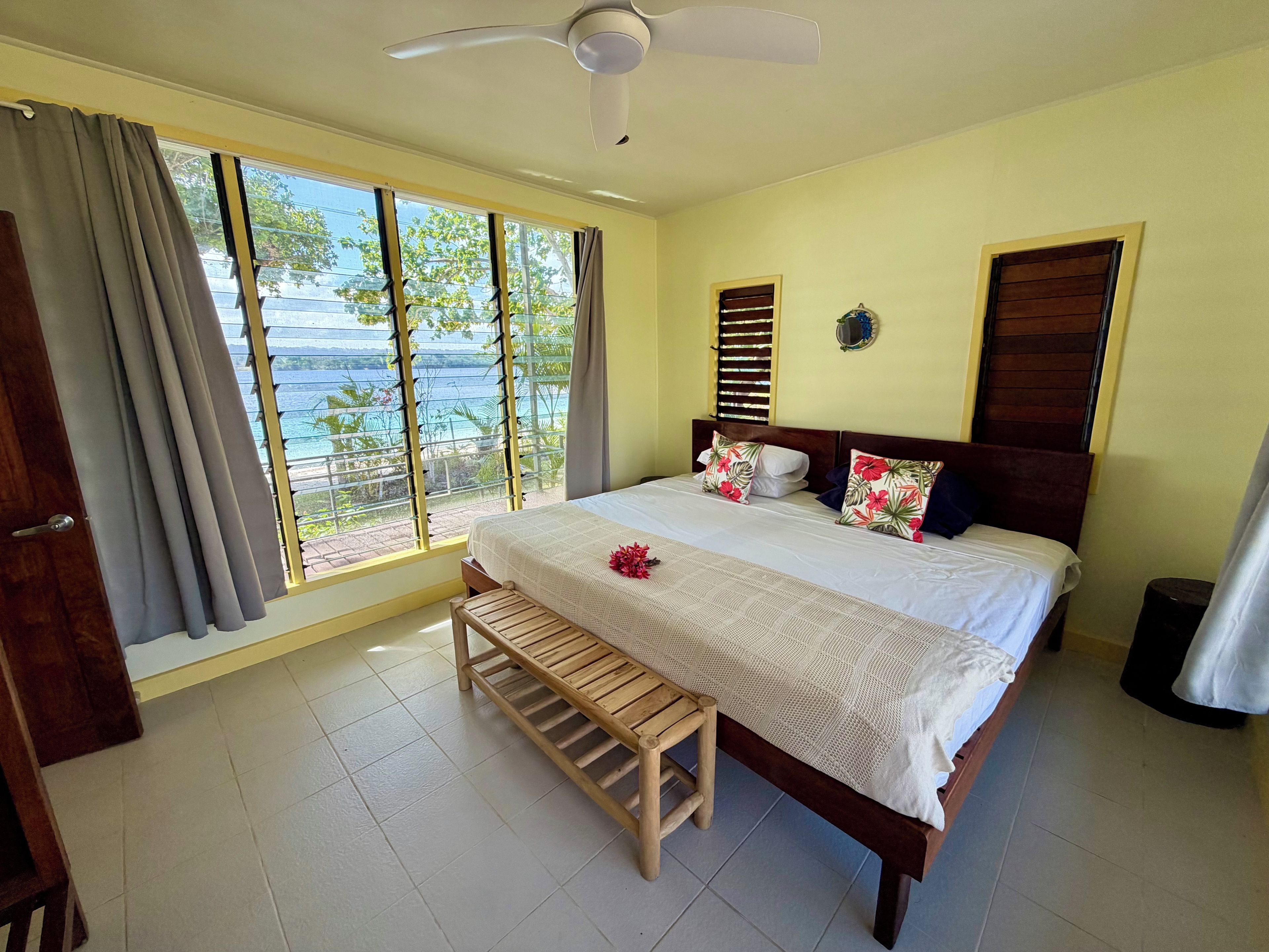 A bedroom with a bed, a wooden bench, a ceiling fan, and a view of the sea.