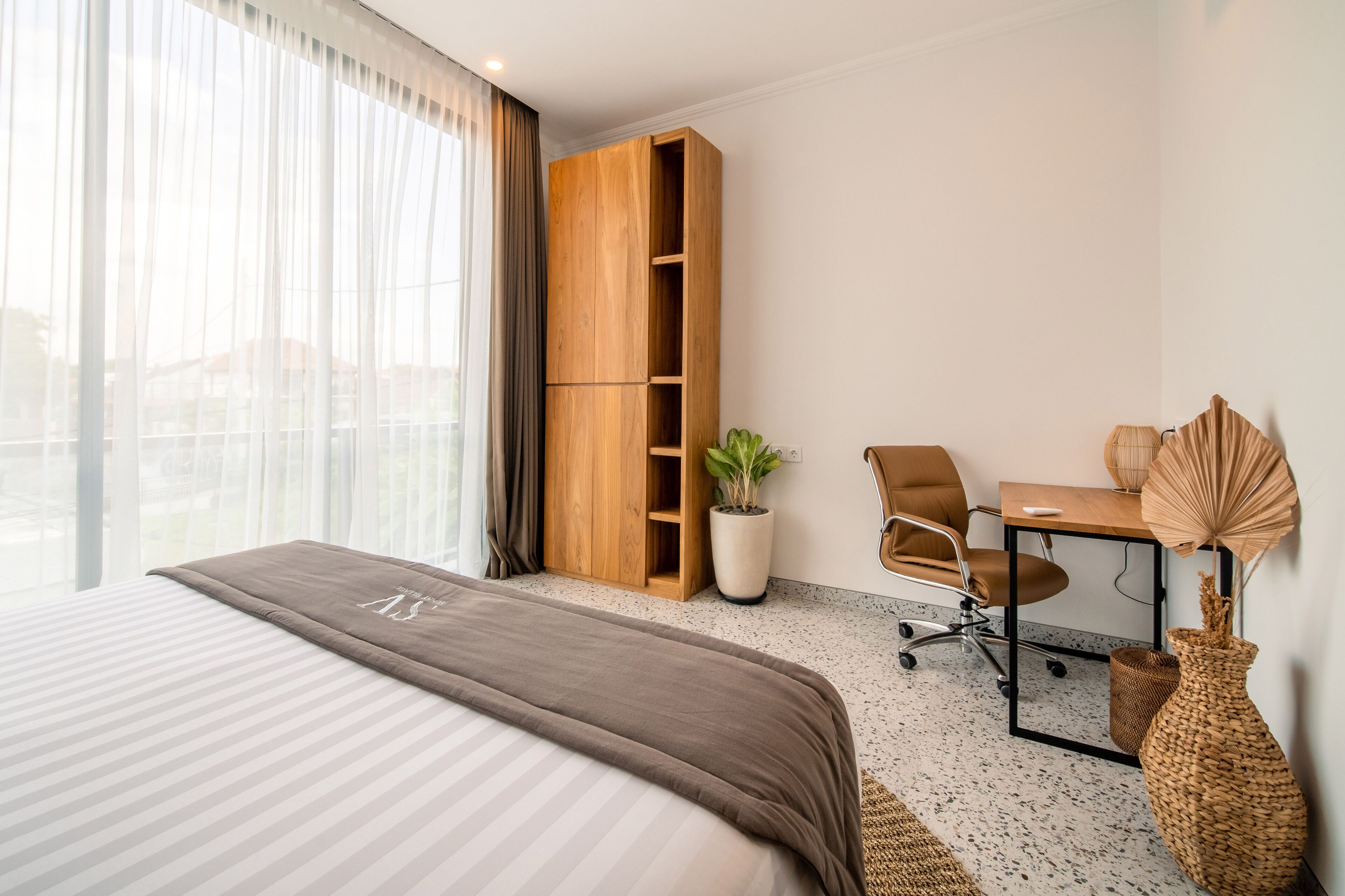 A hotel room with a large bed, a wooden wardrobe, a desk with a chair, a potted plant, and a decorative vase.