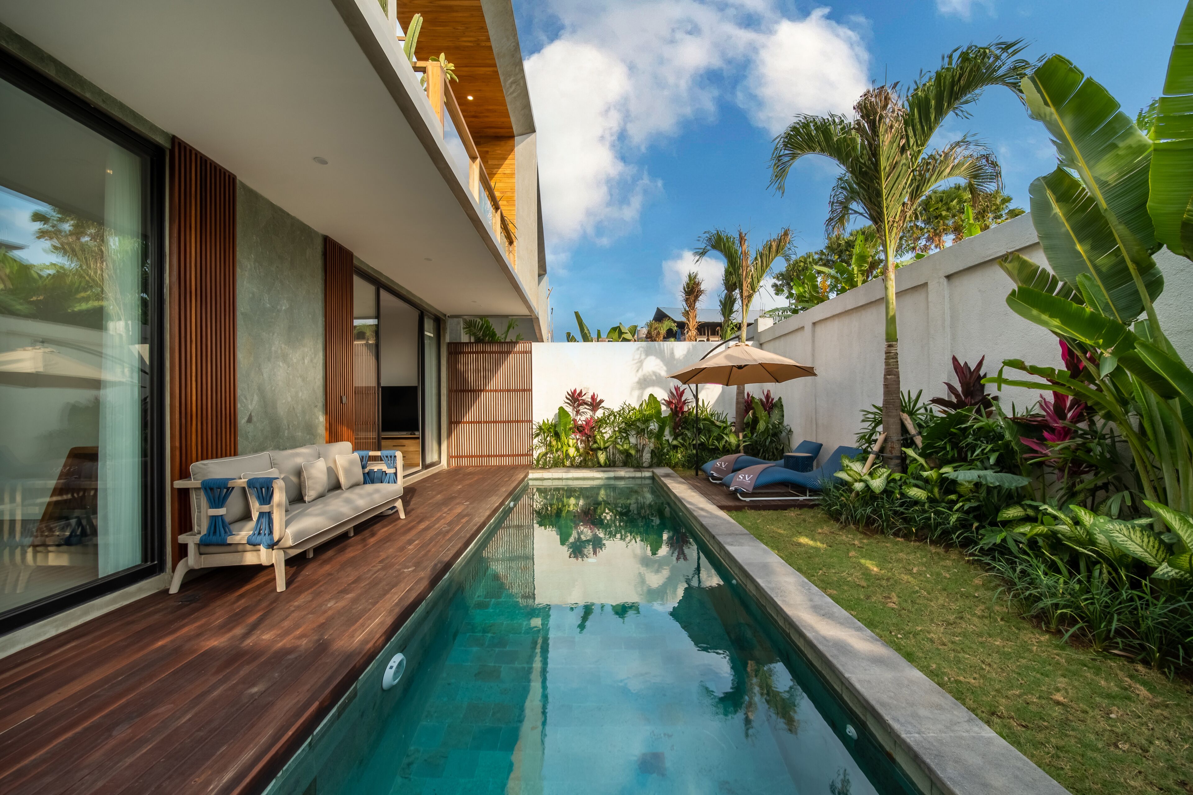 Un espace piscine extérieur moderne, doté d’une terrasse en bois, de chaises longues et de diverses plantes tropicales.