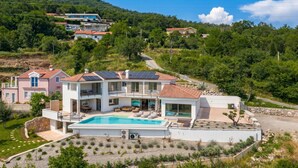 Une maison moderne à deux étages, dotée d’une piscine, entourée de verdure et de collines.