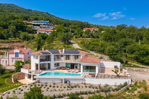 Une maison moderne à deux étages, dotée d’une piscine, entourée d’un jardin aménagé et d’un paysage vallonné.