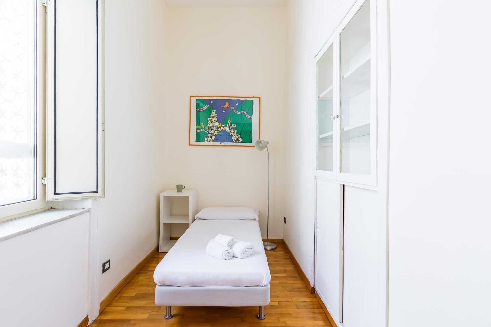 Una habitación pequeña y estrecha con una cama individual, una mesita de noche, una lámpara y un mapa enmarcado en la pared.
