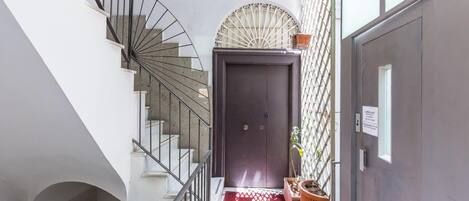 Un pasillo estrecho con una escalera de caracol a la izquierda, una puerta oscura al final y un felpudo rojo en la entrada.