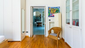 Ein Flur mit Holzboden, einem Korbstuhl, einem weißen Schrank mit Glastüren und einem farbenfrohen Gemälde an der Wand.