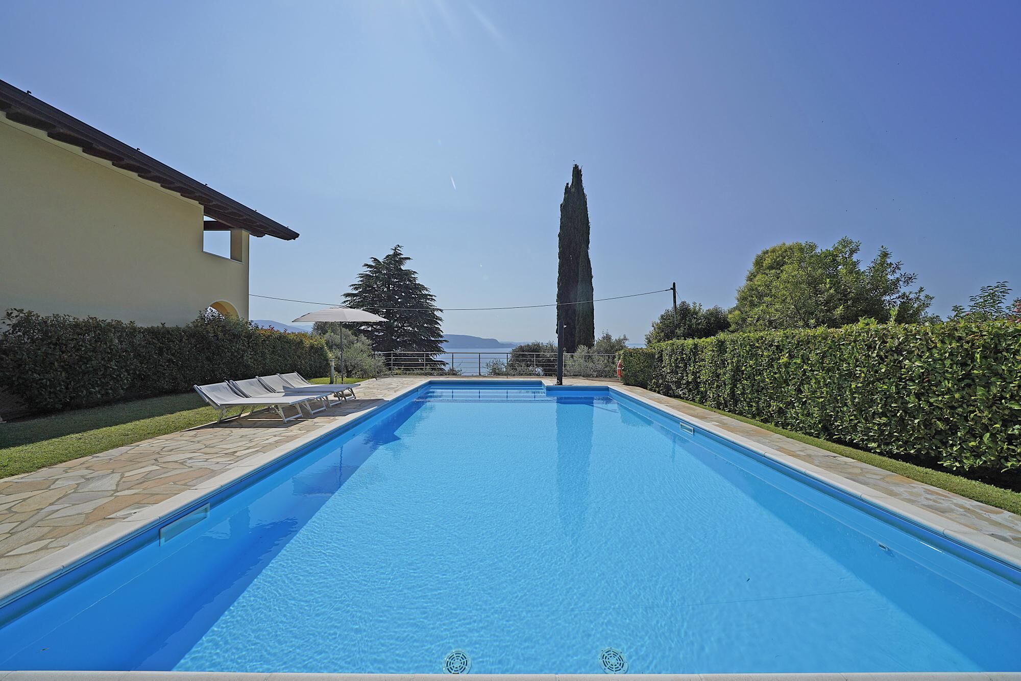 A rectangular swimming pool with a clear blue water surface, surrounded by a stone deck and a hedge.