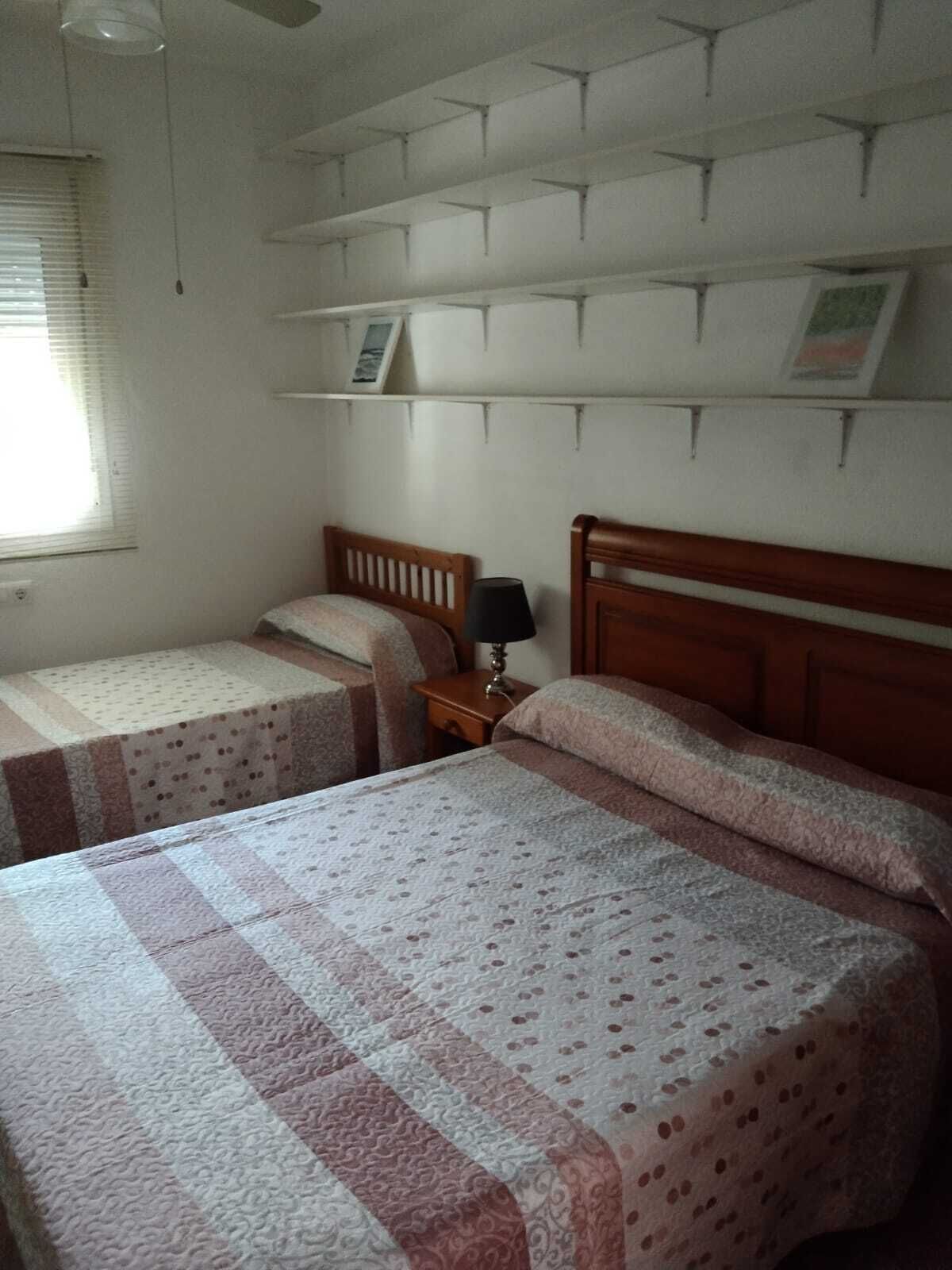 Un dormitorio con dos camas individuales, una mesita de noche con una lámpara y una estantería con marcos de fotos.