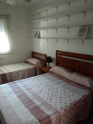 Un dormitorio con dos camas individuales, una mesita de noche con una lámpara y una estantería con marcos de fotos.