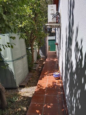 Un estrecho sendero al aire libre que lleva a una verja verde, flanqueado por una pared blanca y un árbol.