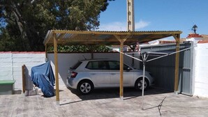 Un coche estacionado bajo una pérgola de madera en un área de estacionamiento techada, con una pared blanca y un árbol al fondo.