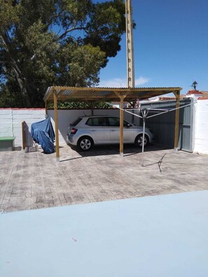 Ein Auto steht unter einer Holzpergola in einer überdachten Parkgarage, im Hintergrund eine Mauer und ein Baum.