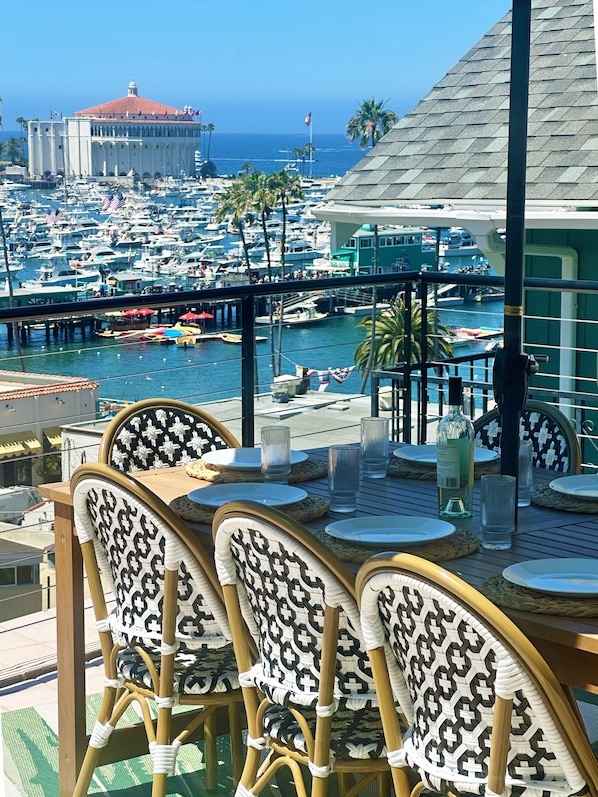 A dining area with wicker chairs and a table set for two, overlooking a marina with boats and a distant building.