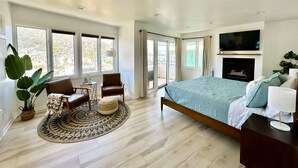 A spacious bedroom with a large bed, a TV mounted above the fireplace, and a view of the city through large windows.