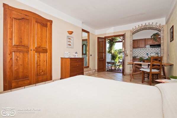 A bedroom with a large wooden wardrobe, a small desk with a chair, and a view of the garden through an open door.