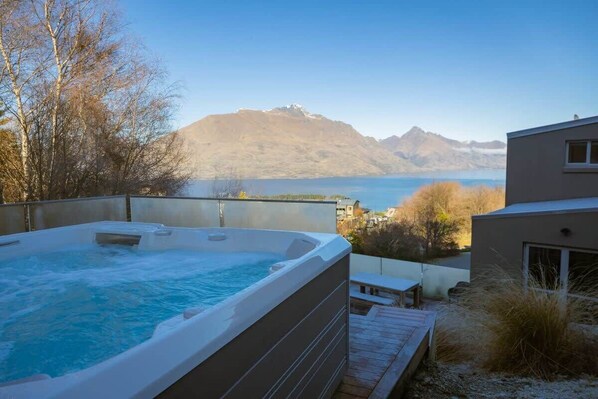 A hot tub with a mountainous backdrop, a wooden deck, and a modern building.