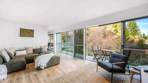A modern living room with a sectional sofa, a coffee table, and a dining area with chairs. Large windows provide a view of the outdoors.
