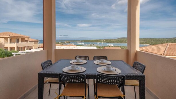 A balcony with a table set for two, overlooking a coastal landscape with buildings and boats.