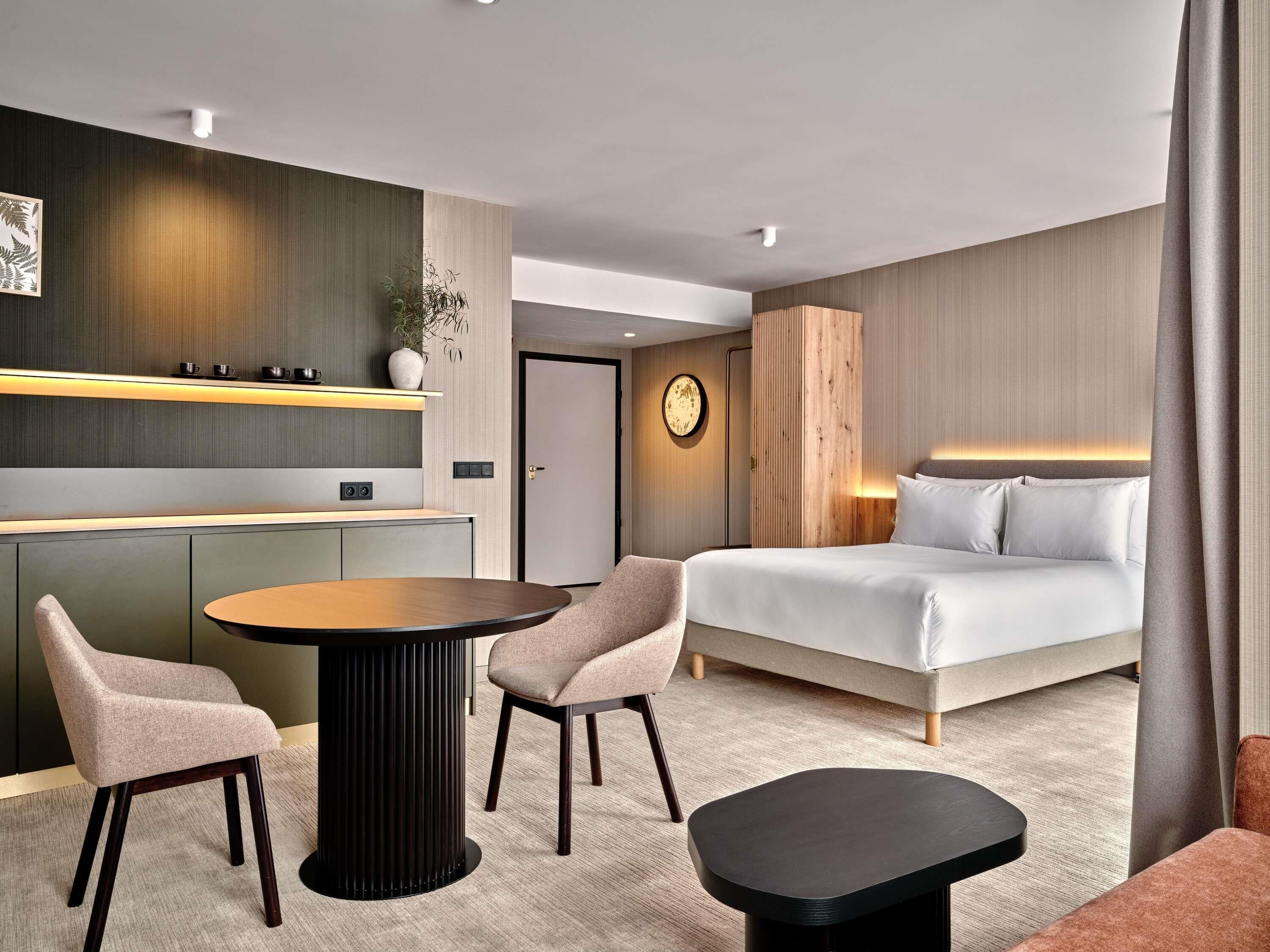 Een moderne hotelkamer met een bed, een ronde eettafel, twee stoelen, een klein zwart nachtkastje, een wandklok en een plant aan de muur.