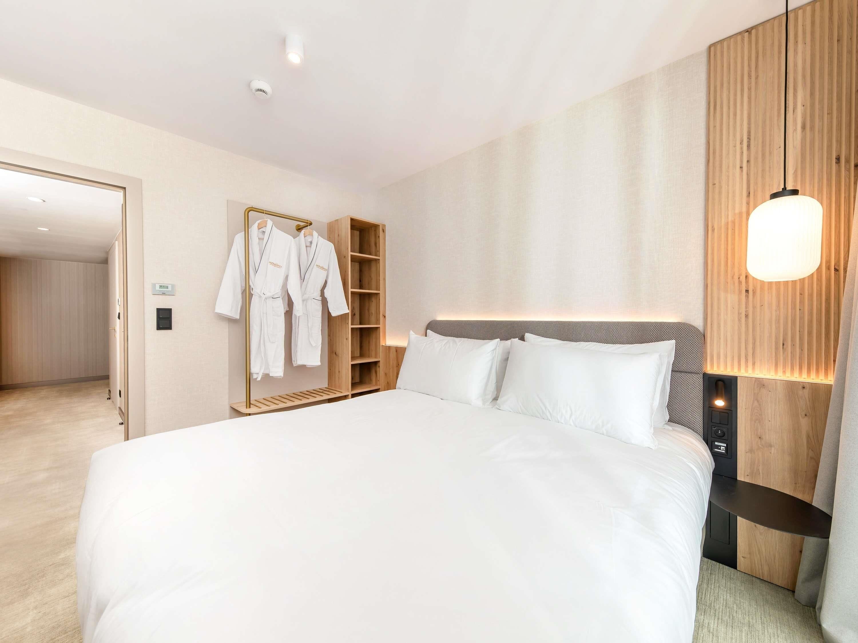 A modern hotel room with a large bed, wooden headboard, and a built-in wardrobe with hanging robes.