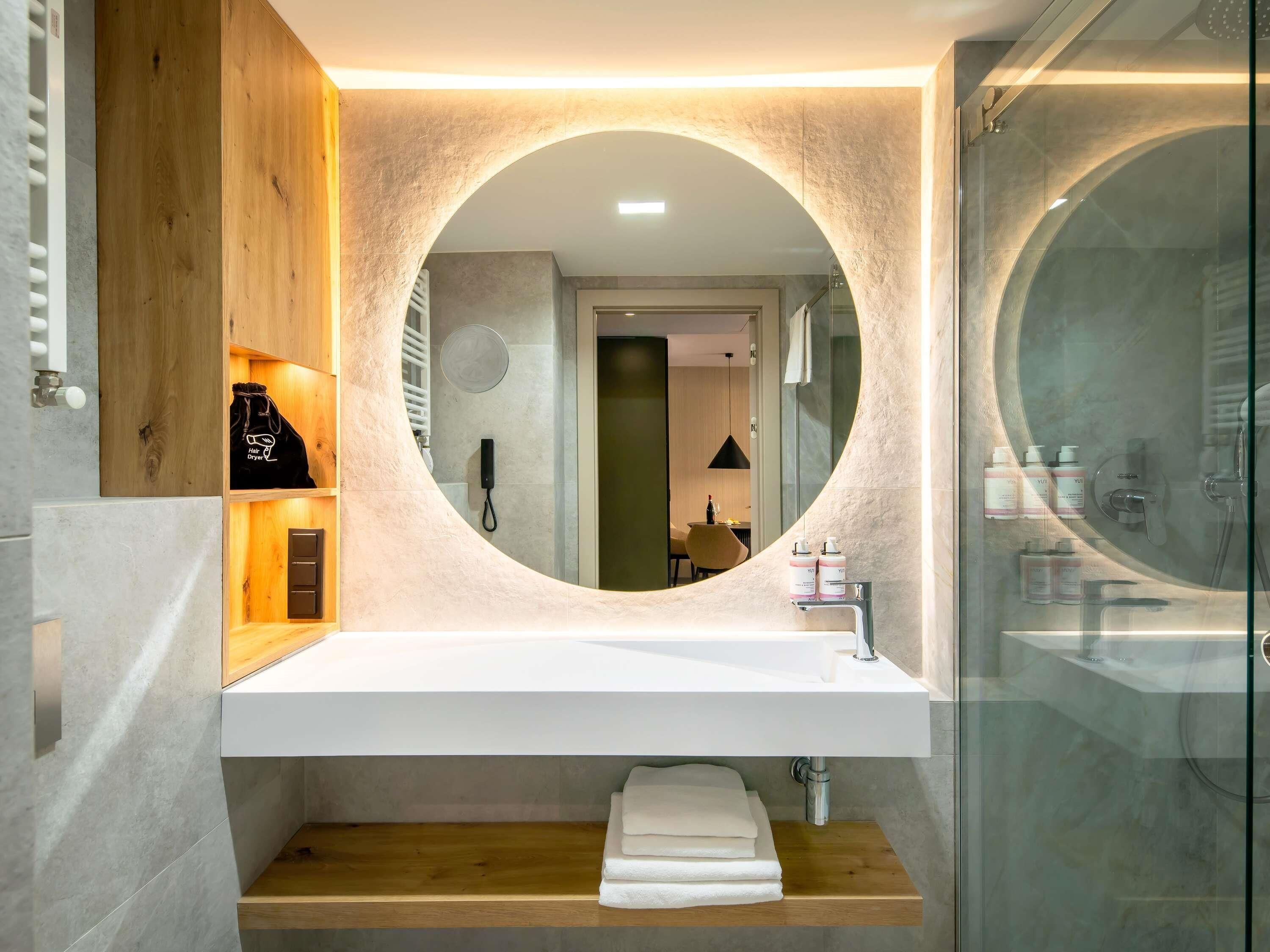 Un baño moderno con un espejo redondo, un lavabo blanco y una ducha con mampara de vidrio.