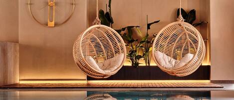 A modern indoor pool area with two wicker hanging chairs, a wall-mounted clock, and a plant arrangement.