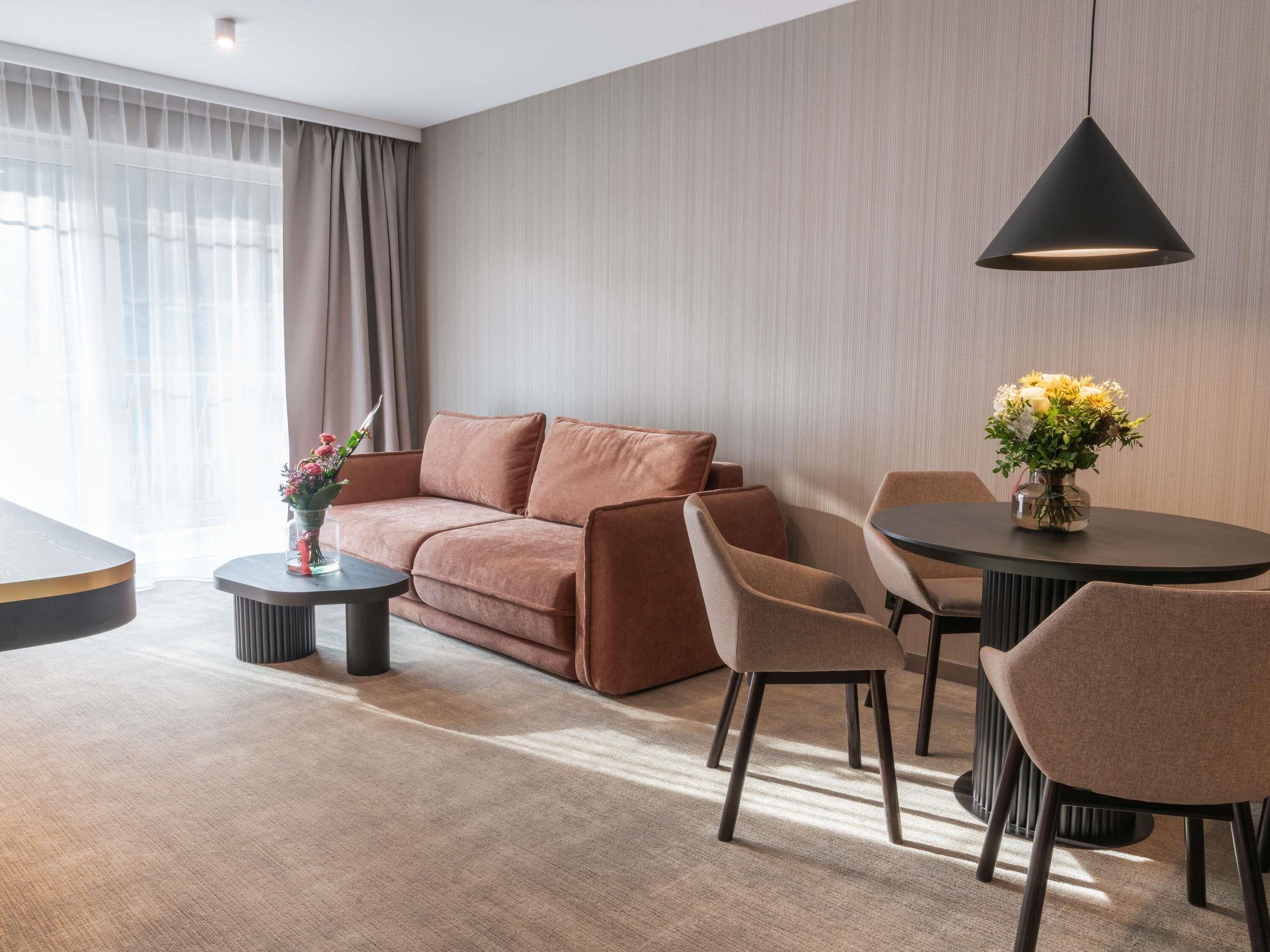 A modern hotel room with a sofa, chairs, a round table, and a pendant light.
