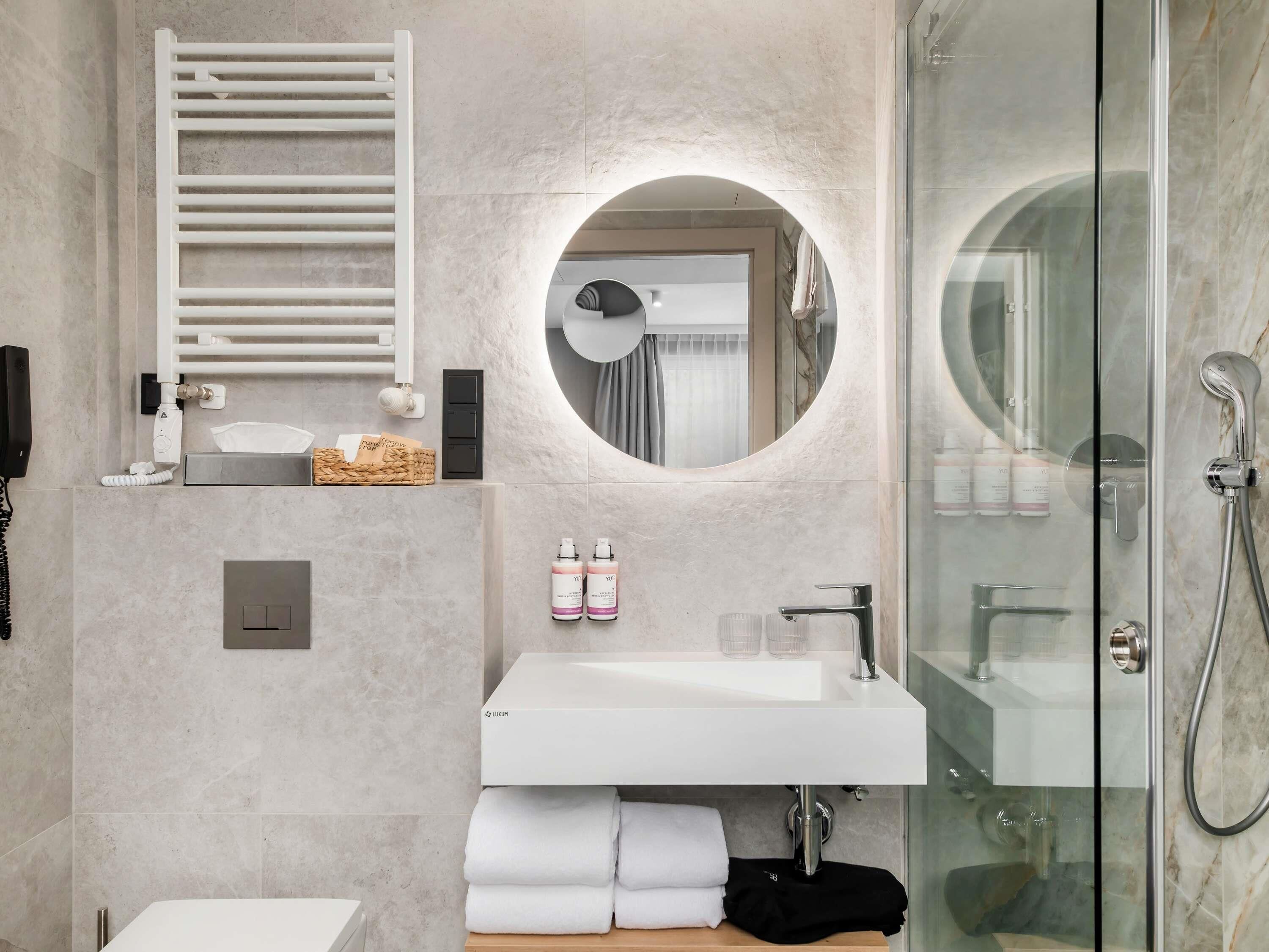A modern bathroom with a glass-enclosed shower, a white sink, and a round mirror.