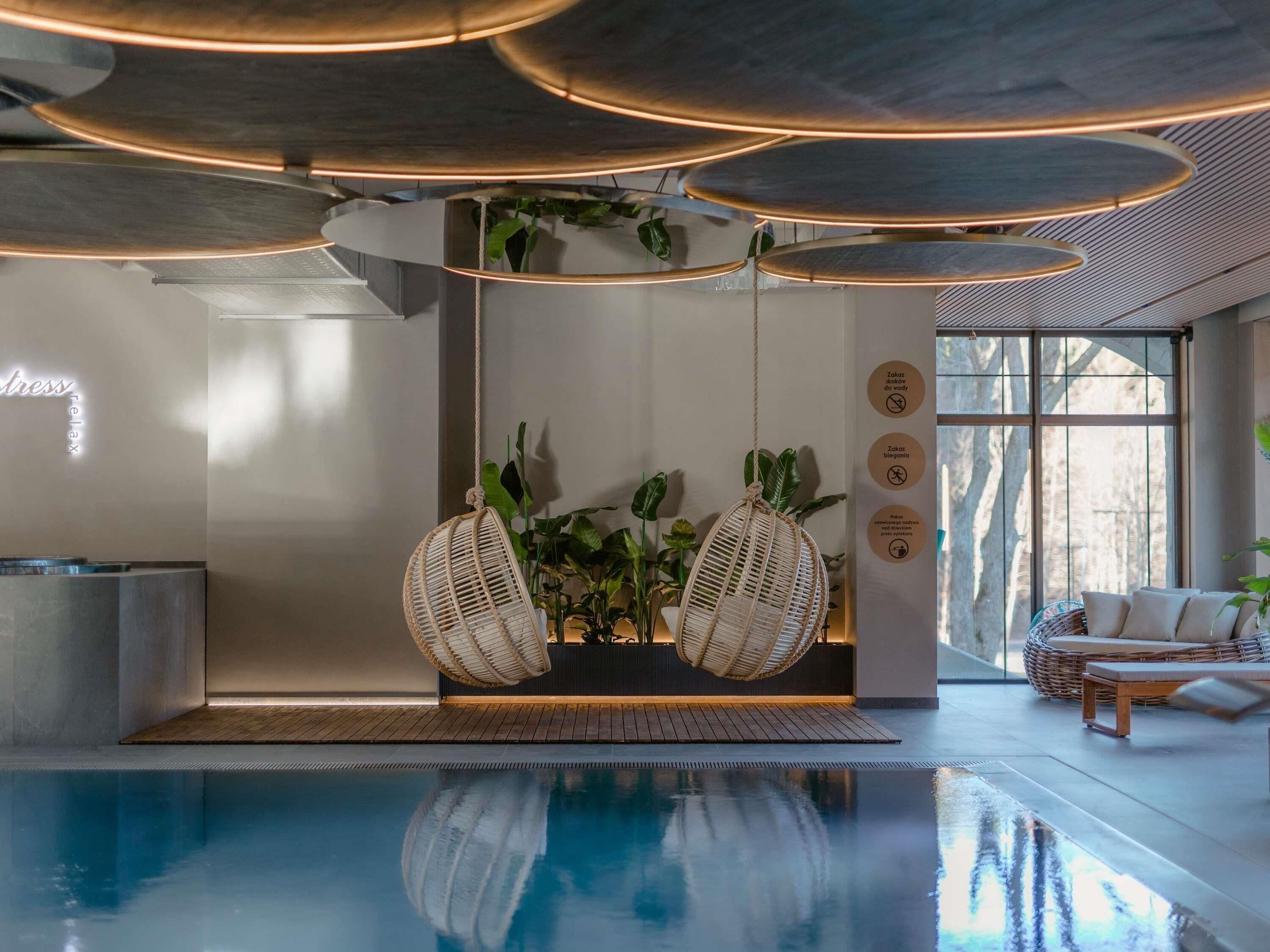 Un espace piscine intérieur moderne, comprenant un comptoir d’accueil, deux fauteuils en osier et une grande fenêtre.