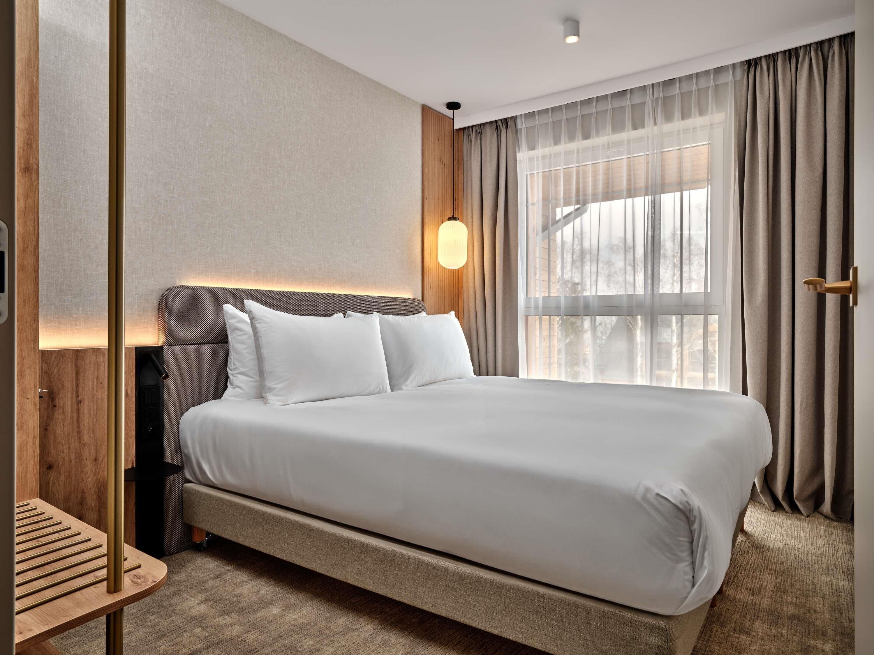 A modern hotel room with a large bed, a wooden headboard, a bedside table, and a window with curtains.