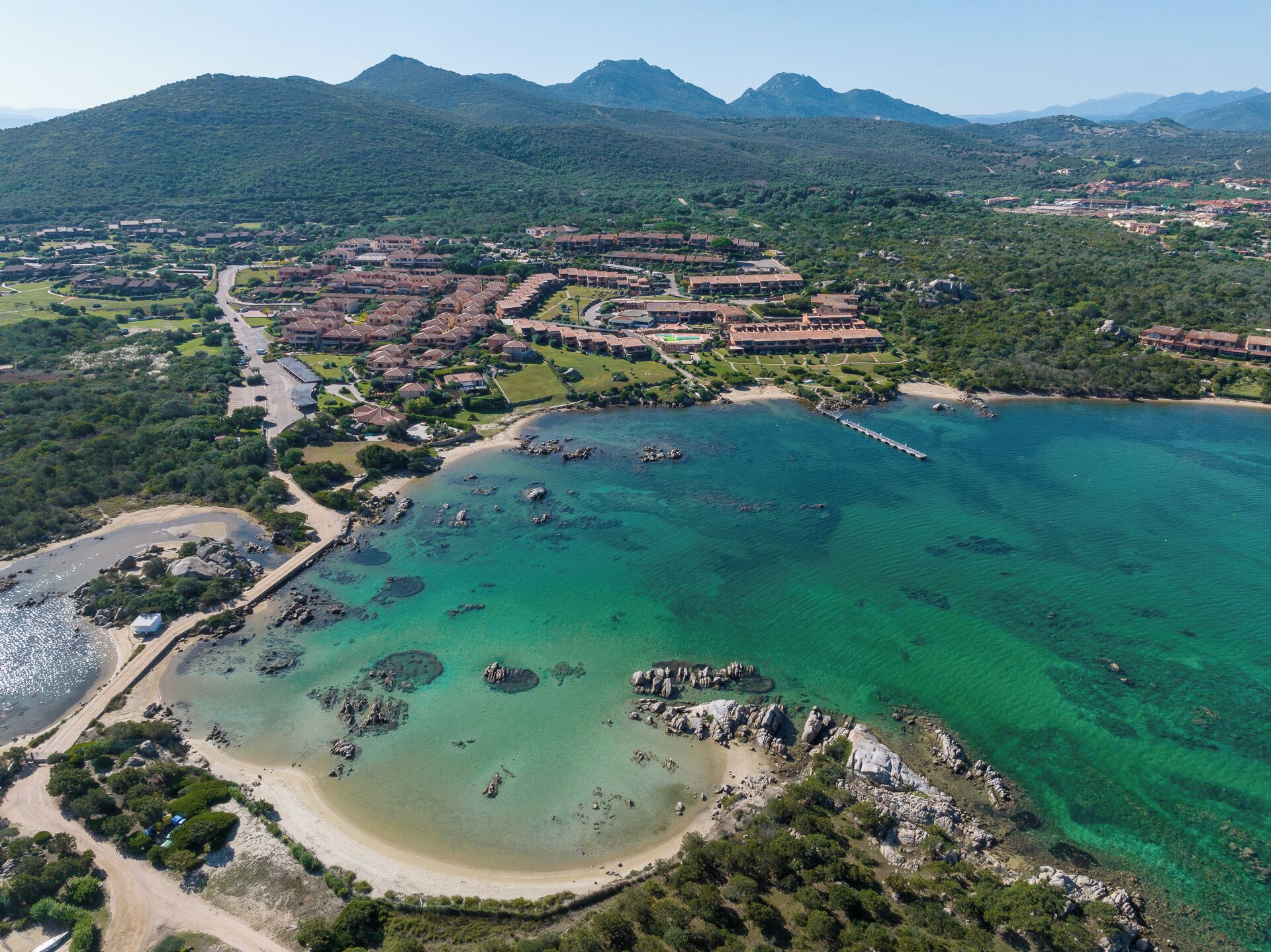 Luftaufnahme eines Küstenresorts mit mehreren Gebäuden, einem Strand und klarem türkisblauem Wasser.