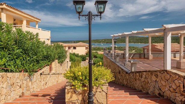 Una escalera de piedra con una farola, que lleva a un espacio exterior cubierto con vista al mar y edificios.