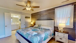 Une chambre à coucher comprenant un lit, une table de chevet, un ventilateur de plafond, une fenêtre avec des rideaux et une porte donnant sur une autre pièce.