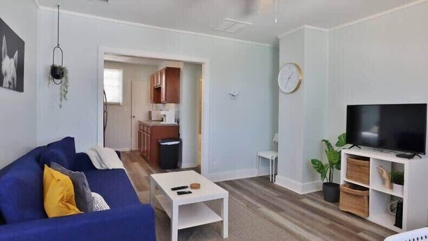 Ein Wohnzimmer mit einem blauen Sofa, einem Couchtisch, einem Fernseher auf einem weißen Schrank und einer Küche im Hintergrund.