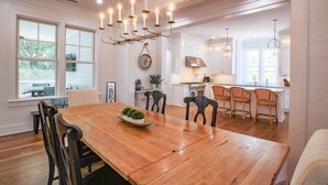 A spacious dining area with a wooden table and chairs, a chandelier, and a view of the kitchen and dining room in the background. - Upscale, Custom Home 3 Doors From Beach w/ Private Pool, Hot Tub & Screened Porch, Bunk Room & Den (Isle of Palms)
