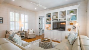 A spacious living room with a beige sofa, a coffee table, and a TV on a white cabinet. - Upscale, Custom Home 3 Doors From Beach w/ Private Pool, Hot Tub & Screened Porch, Bunk Room & Den (Isle of Palms)