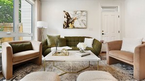A modern living room with a green sofa, beige armchairs, and a white coffee table. There is a painting on the wall and a window with blinds.