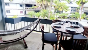 A balcony with a hammock, a table set for dining, and a view of a resort with buildings and palm trees.