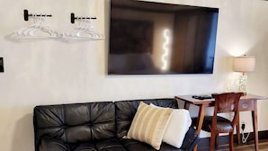 A black leather sofa with a white and beige pillow, a wooden dining table with chairs, and a flat-screen TV mounted on the wall.