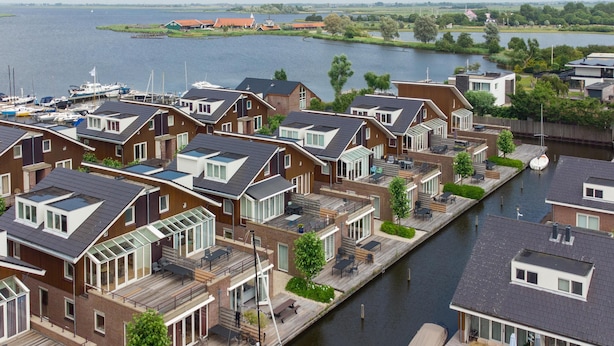 A canal-side residential area with modern houses featuring large windows and balconies, a dock with a boat, and a view of the water and distant landscape. - luxury apartment with waterside terrace (Uitgeest)