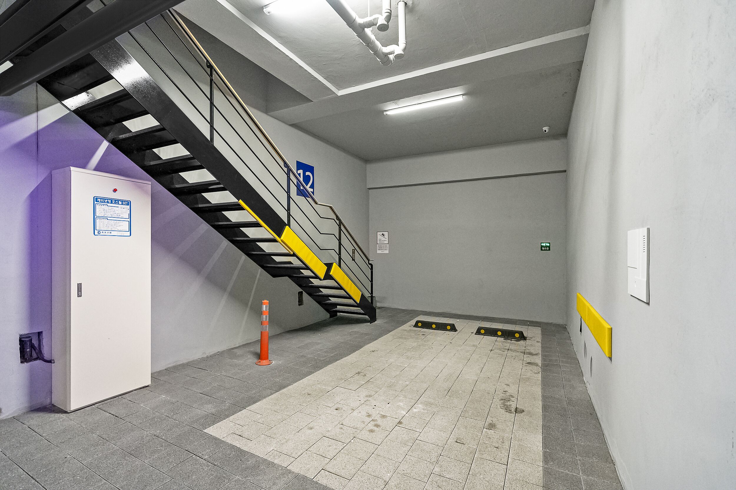 A stairwell with a handrail, a sign indicating room number 12, a fire alarm box, and a safety cone.