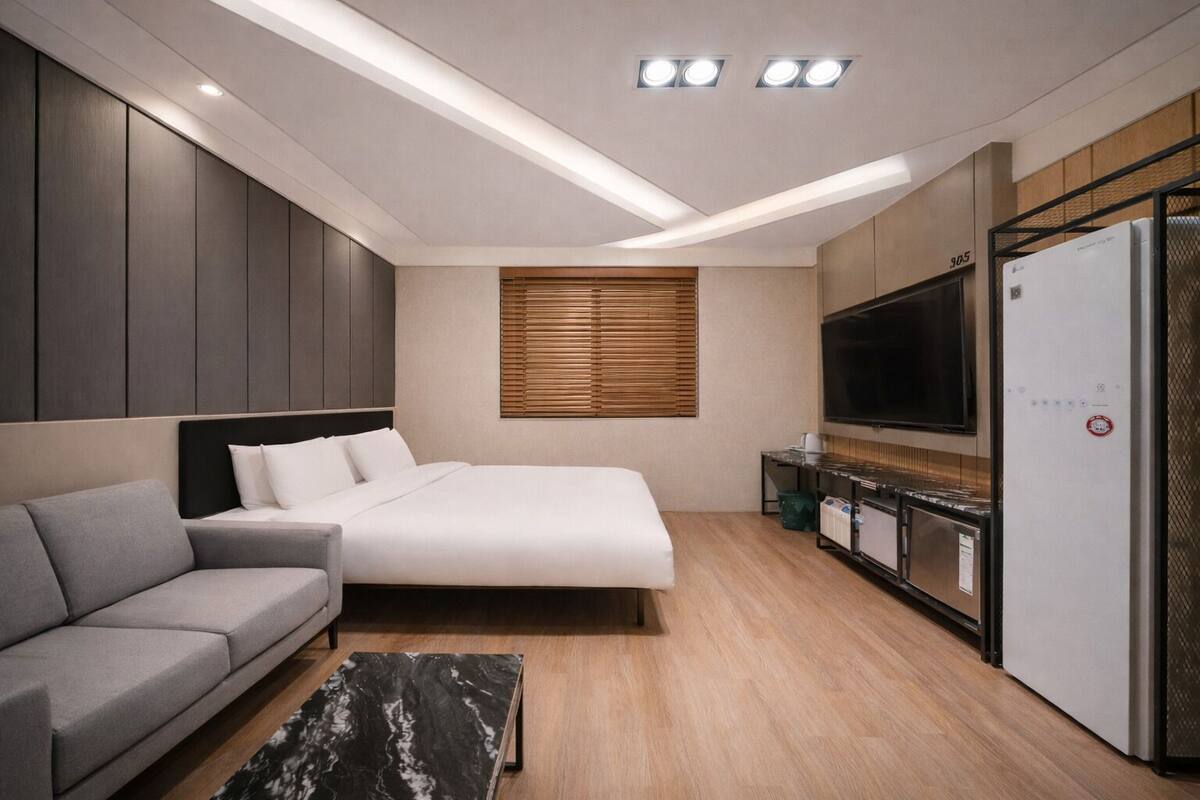 A modern hotel room with a bed, sofa, TV, kitchenette, and wooden flooring.