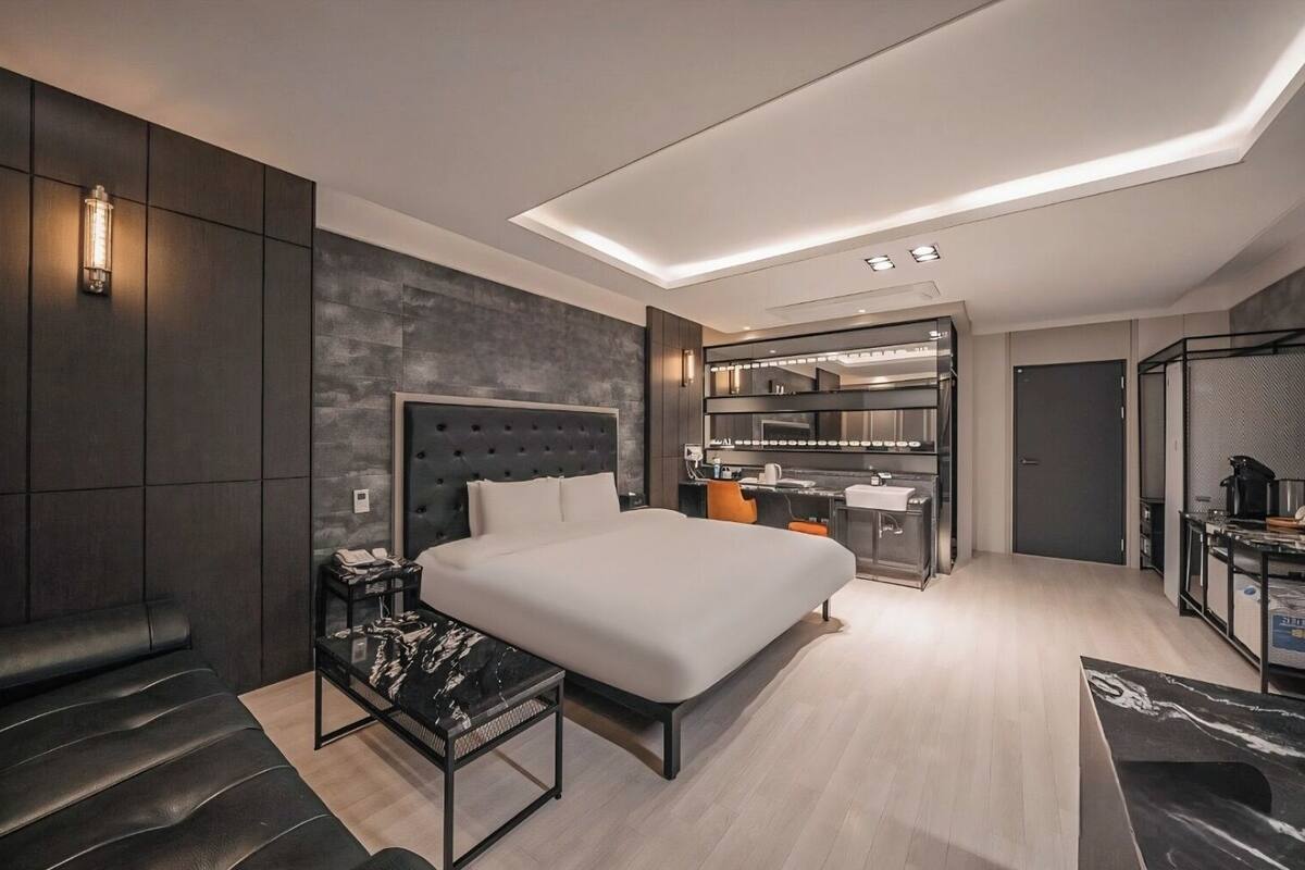 A modern hotel room with a large bed, a desk, a seating area, and a kitchenette.