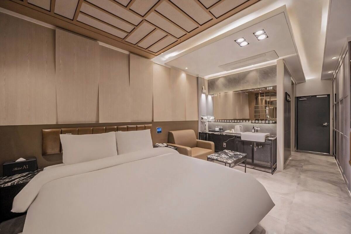 A modern hotel room with a large bed, a desk, a chair, a minibar, and a bathroom with a mirror and sink.