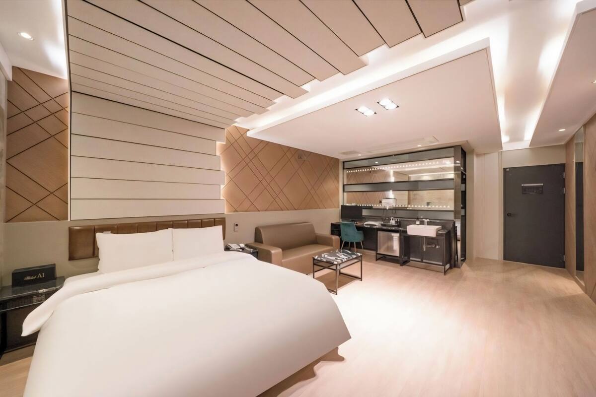 A modern hotel room with a large bed, a sofa, a desk, and a kitchenette.