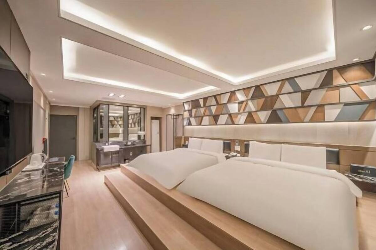 A modern hotel room with a large bed, a desk with a chair, a vanity with a mirror, and a geometric-patterned wall.